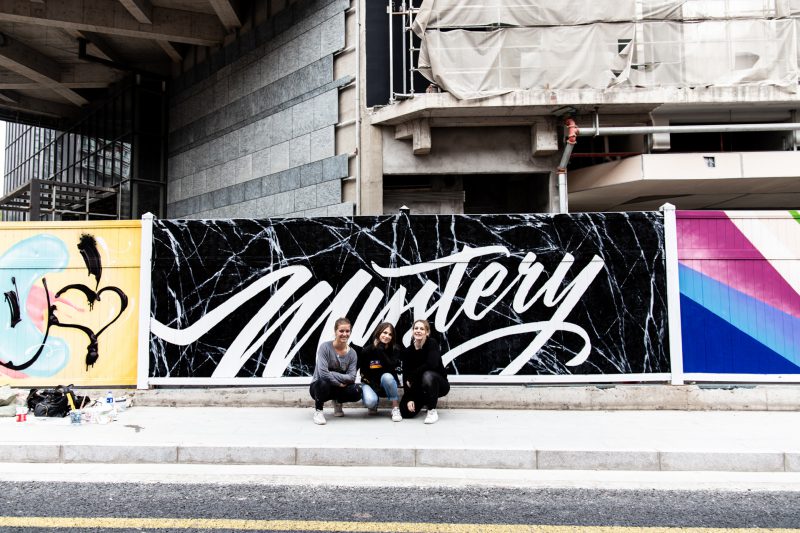 Three women crouching in front of a mural of the word mystery.